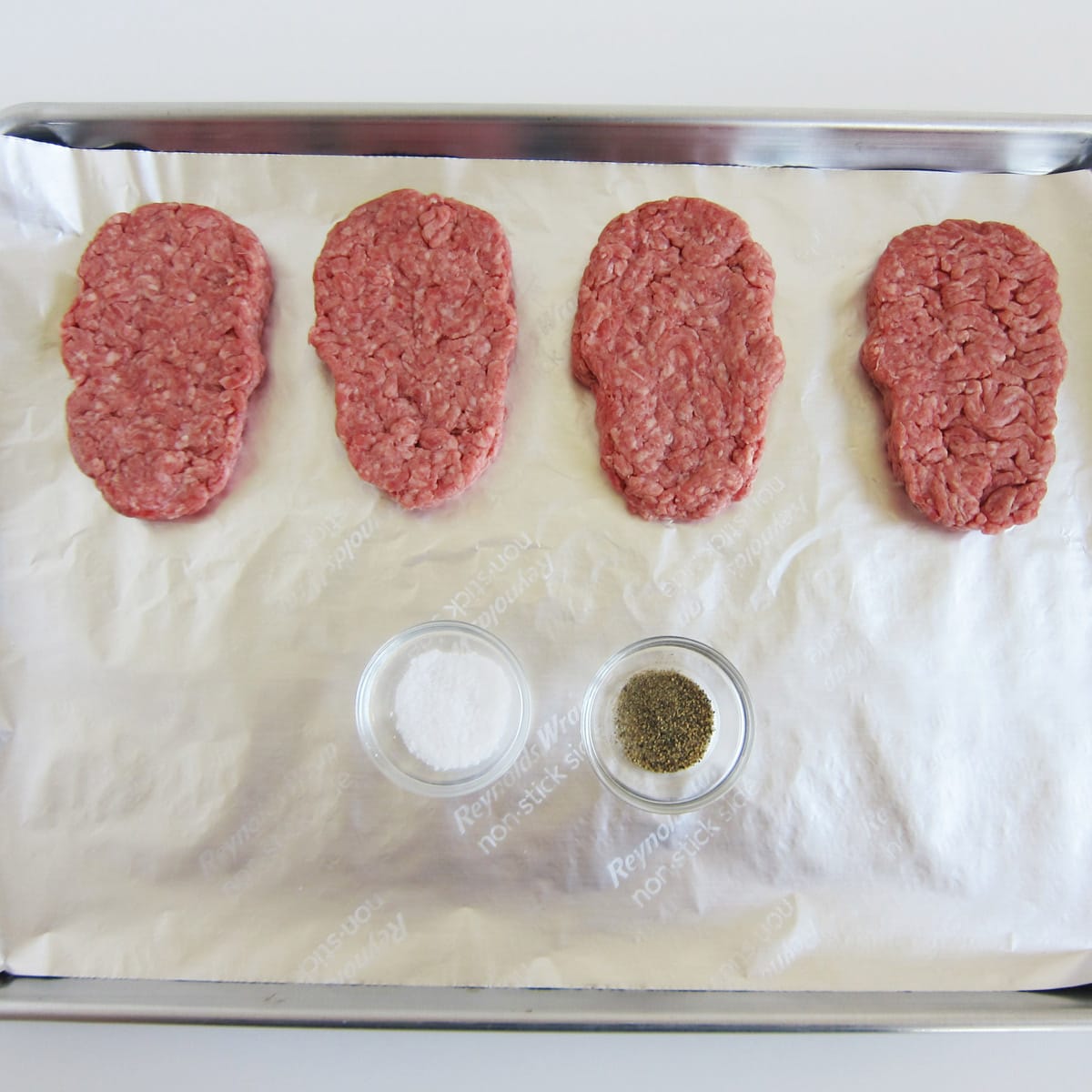 uncooked skull-shaped hamburger patties on a tin foil-lined baking tray with salt and pepper.