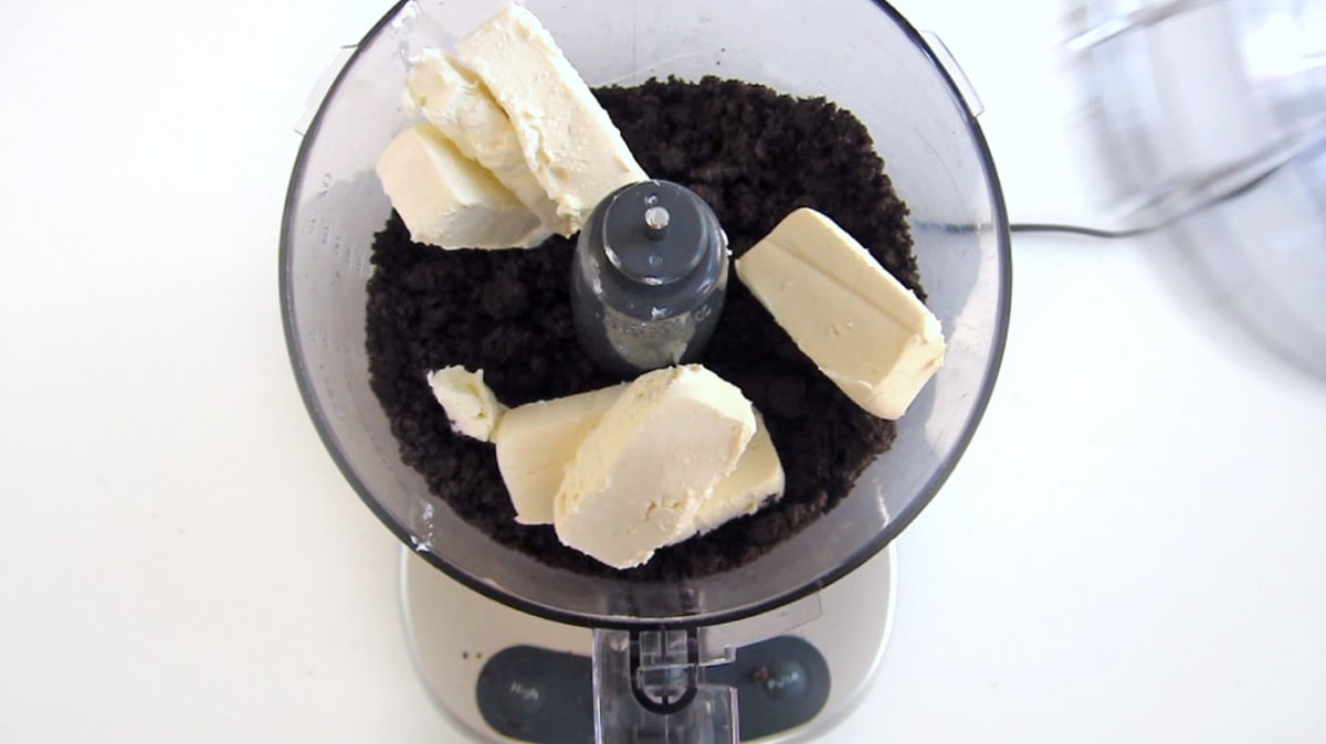 cream cheese added to a food processor bowl filled with OREO Cookie crumbs.