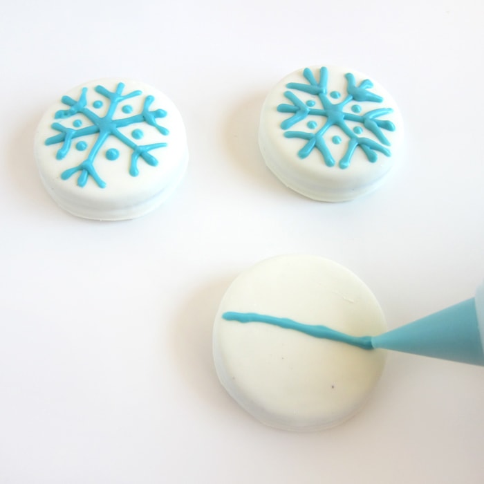 piping blue candy melts onto white fudge covered OREOs to add a snowflake design.